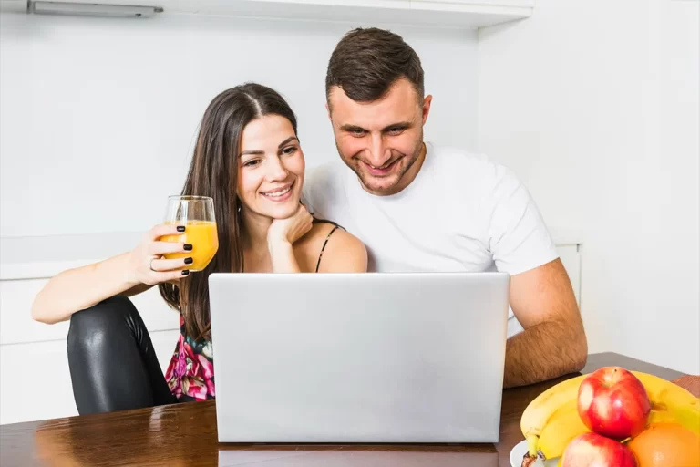 Mutfak masasında laptop başında oturan, meyve sularını yudumlarken gülümseyerek online BES başvurusu işlemlerini gerçekleştiren genç çift.