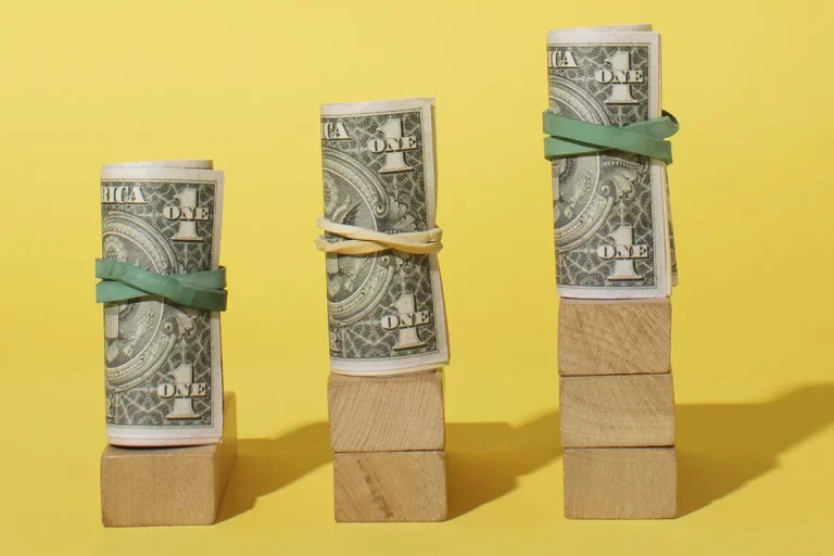 Sarı zemin üzerinde ahşap bloklar üzerinde yükselen dolar ruloları ve dovize endeksli doviz bazli hayat sigortasi ile enflasyona karşı koruma sağlayan yatırım planı.